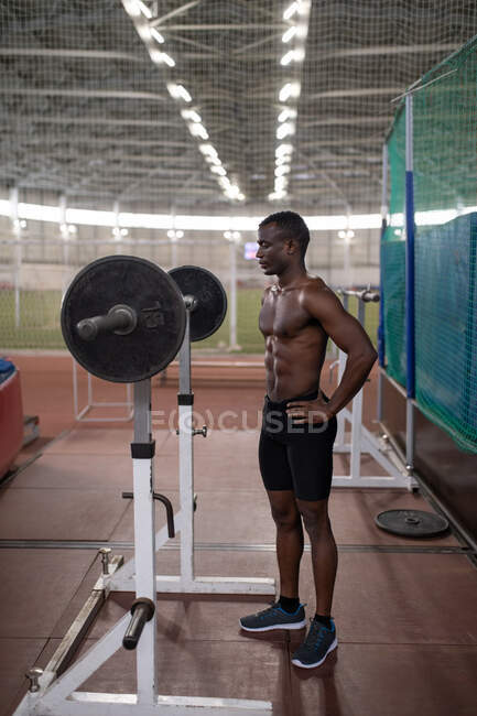 Афроамериканский спортсмен с руками на талии готовится поднять штангу на стадионе — стоковое фото