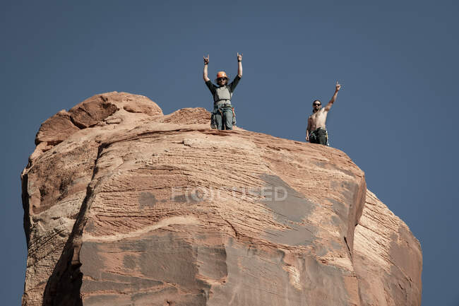 Низкий угол обзора друзей-мужчин, стоящих с поднятыми руками на вершине горной породы на фоне ясного неба — стоковое фото