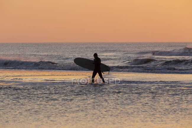 Un surfista caminando por la orilla al atardecer - foto de stock