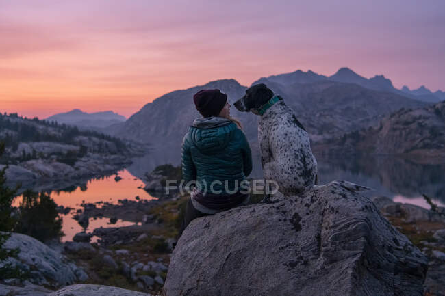 Vista posteriore di giovane donna seduta su roccia con cane in montagna durante il tramonto — Foto stock
