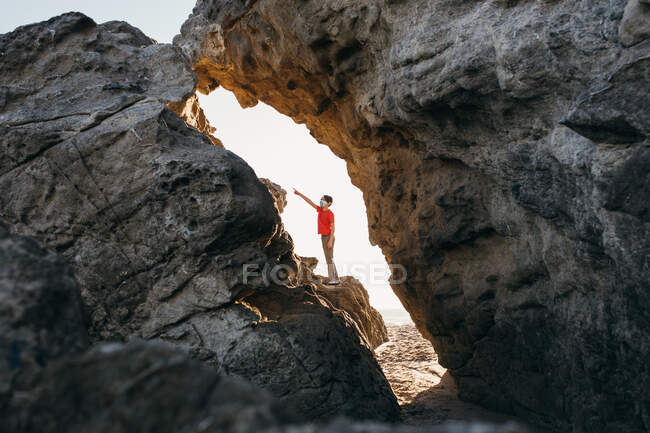 Garçon portant une chemise rouge et masque visage points sur les rochers à la plage — Photo de stock
