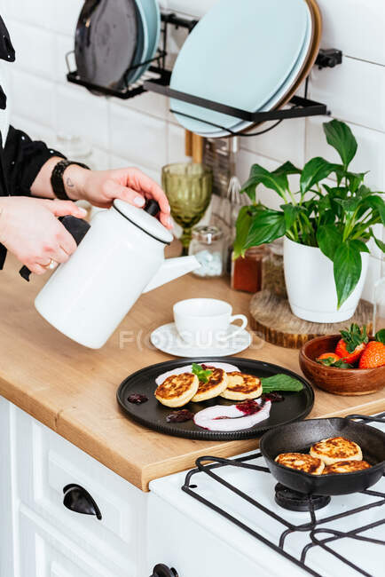 Les mains des femmes versent le thé dans une tasse. crêpes au fromage avec confiture sur une assiette noire — Photo de stock