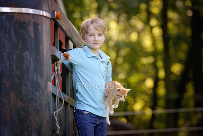Guapo chico rubio sosteniendo un gatito al aire libre en un entorno rural. - foto de stock