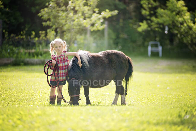 Cute little blond girl with arm around black pony. — Stock Photo