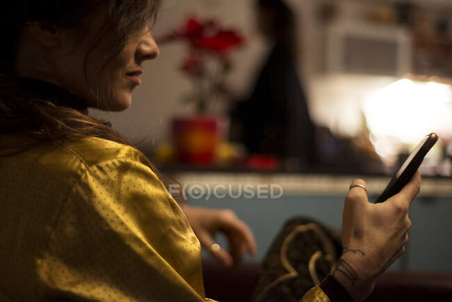 Frau im goldenen Seidenhemd zu Hause kuschelige Küche blickt auf Handy-Gerät — Stockfoto