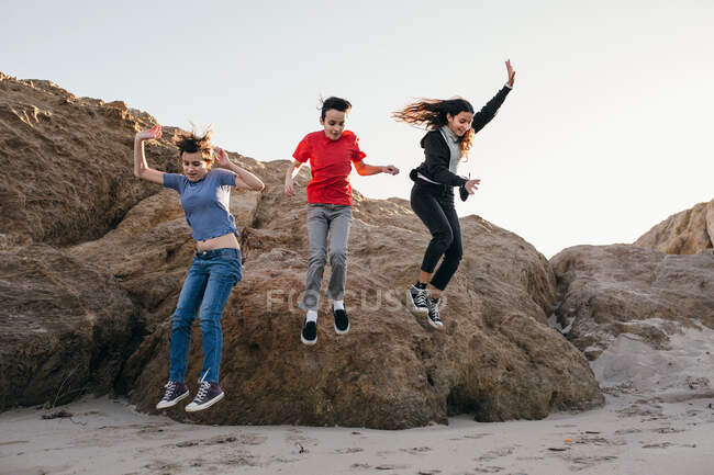Drei geschwister springen von einem großen felsen am strand — Stockfoto