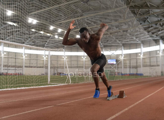 Muscular black athlete running fast from starting block in track and field stadium — Fotografia de Stock