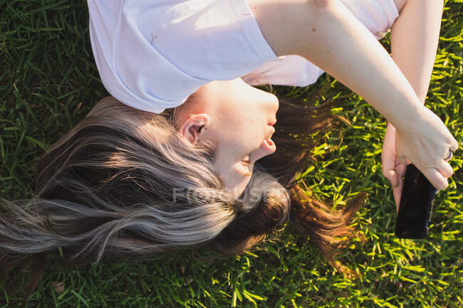 Retrato de una chica tumbada en la hierba con un teléfono en las manos al atardecer - foto de stock