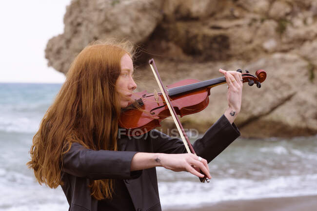 Молода руда жіноча скрипалька грає на скрипці на пляжі в похмурий день — стокове фото