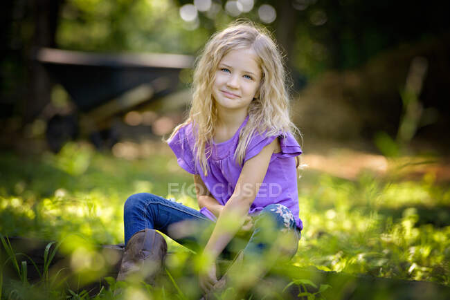 Linda menina loira em camisa roxa sentado na grama. — Fotografia de Stock
