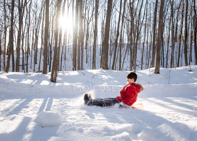 Kleines Kind rodelt an sonnigem Tag einen verschneiten Hügel in einem Waldgebiet hinunter. — Stockfoto