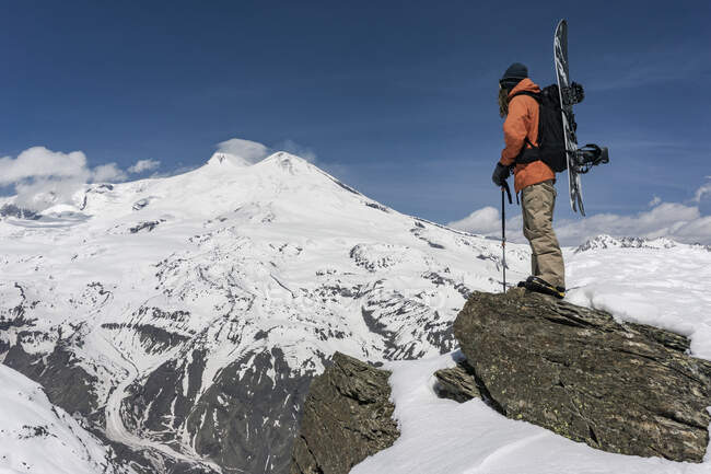 Homem com bastão de esqui e snowboard em pé na rocha na montanha coberta de neve contra o céu — Fotografia de Stock