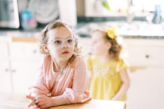 Pequena menina gêmea com óculos olhando sentado na mesa da cozinha — Fotografia de Stock
