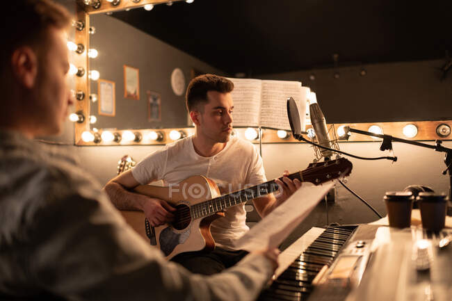Hombre adulto tocando música en la guitarra mientras crea canciones con un amigo en el estudio - foto de stock
