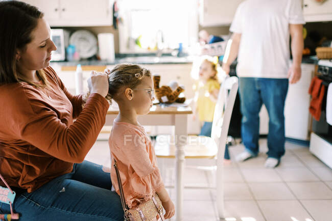 Pequena menina em idade pré-escolar recebendo cabelo feito por sua mãe na cozinha — Fotografia de Stock