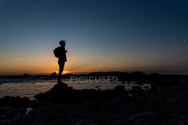 Silhouetted figure standing on rocky shore with sunset sky behind — Stock Photo