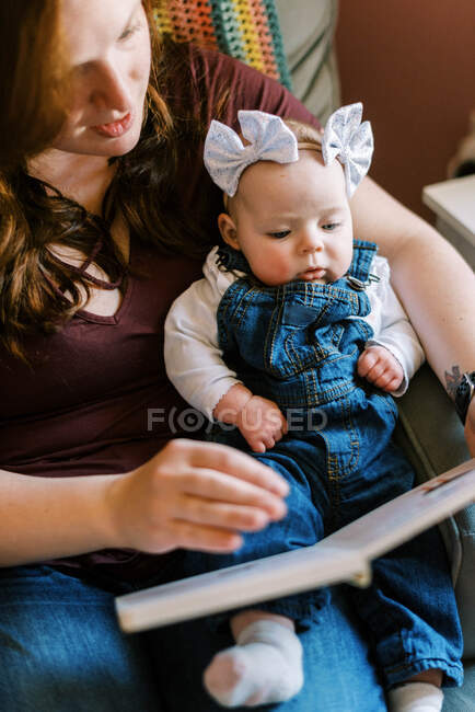 Uma jovem mãe lendo seu bebê um livro na cadeira de balanço do berçário — Fotografia de Stock