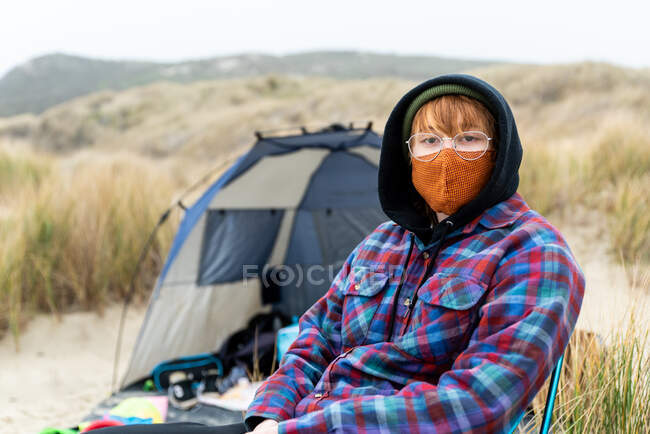 Adolescente en silla de campamento en la costa con máscara mirando a la cámara cerca de las dunas - foto de stock