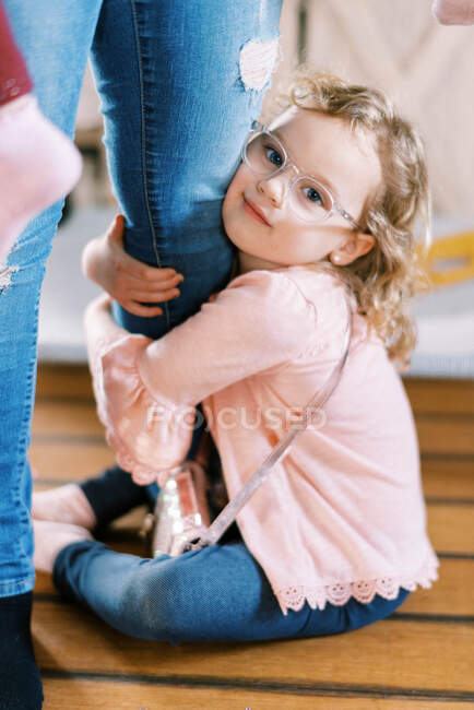 Una bambina con gli occhiali aggrappata alla gamba di sua madre in casa — Foto stock