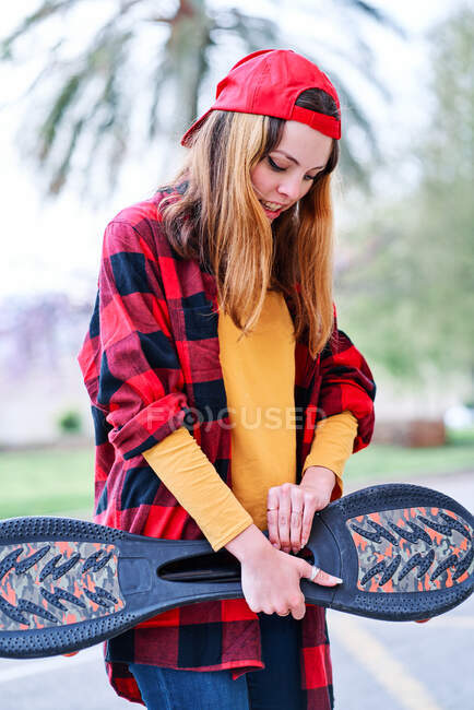 Vertical photo of a young skateboarder girl in plaid shirt and red cap looking down in the city — Stock Photo