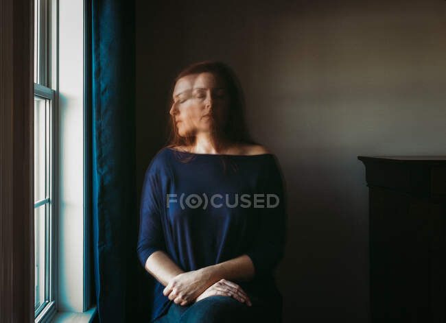 Mujer con la cara borrosa sentada sola en una habitación oscura junto a una ventana. - foto de stock