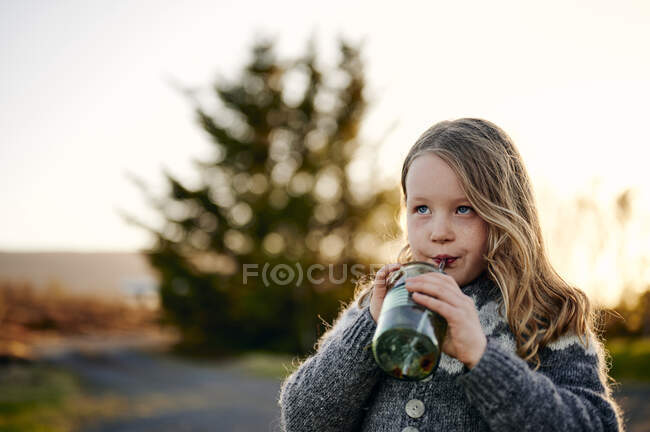 Cute girl sipping fresh drink from jar and looking up while resting in park at sunset — Stock Photo