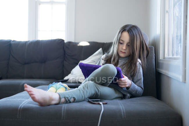 Niña sentada en el sofá usando una tableta. - foto de stock