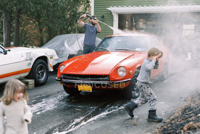 Crianças pequenas ajudando seu pai a lavar um clássico carro vermelho velho lá fora — Fotografia de Stock