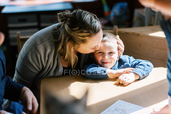 Madre besando a su hijo pequeño sol en la mesa de comedor en la luz de la mañana - foto de stock