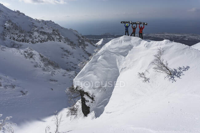 Люди с поднятыми руками держат сноуборды, стоя на заснеженной вершине горы — стоковое фото