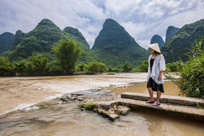 Mulher em chapéu asiático tradicional em pé na margem do rio em Yangshuo — Fotografia de Stock