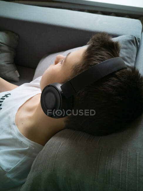 Un niño de 15 años con una camiseta blanca yace en el sofá con auriculares - foto de stock