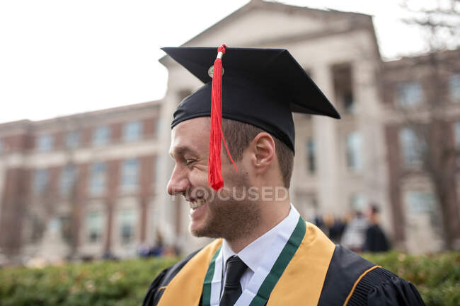 Orgulloso joven sonriendo con alegría en gorra y vestido en la universidad - foto de stock
