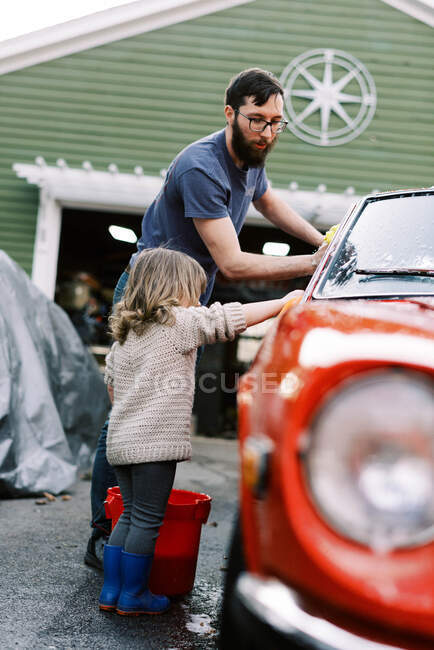 Маленькая девочка моет красную классическую машину вместе со своим отцом — стоковое фото