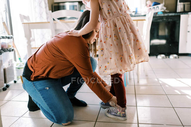 Мама помогает дочери надевать обувь со шнурками на кухне — стоковое фото