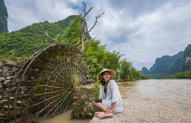 Frau mit traditionellem asiatischen Hut sitzt neben Wassermühle in Yangshuo — Stockfoto