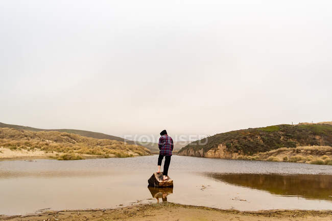 Una persona de pie en el tronco en el borde del agua costera frente a las colinas - foto de stock
