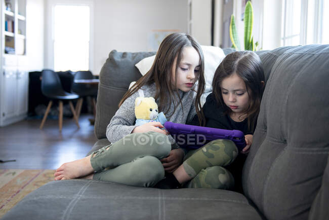 Two little girls sitting on the couch using a tablet. — Stock Photo