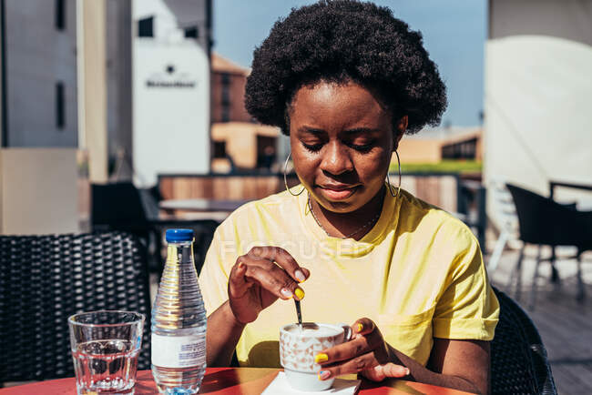 Ritratto di ragazza nera con capelli afro e orecchini a cerchio che beve caffè e una bottiglia d'acqua su una terrazza del bar. — Foto stock