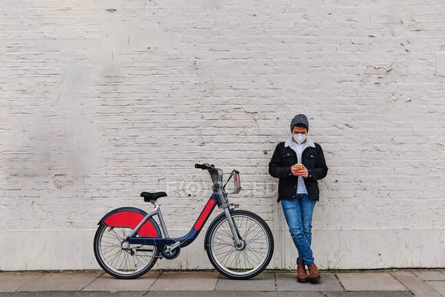 Full body young guy in casual clothes and mask leaning on brick wall near modern bicycle and browsing mobile phone on city street during pandemic in London — Stock Photo