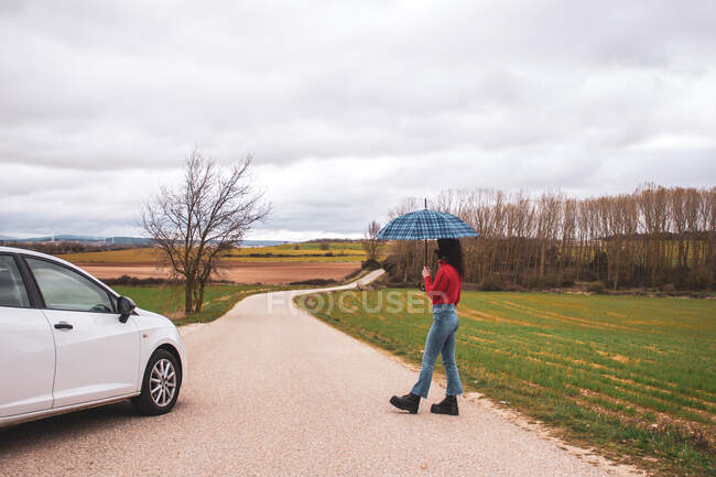 Mujer con paraguas en medio de la carretera junto al campo un coche - foto de stock