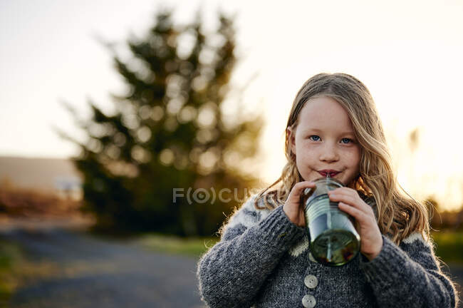 Девушка с длинными волосами пьет свежий напиток из банки и смотрит в камеру во время отдыха в парке на закате — стоковое фото
