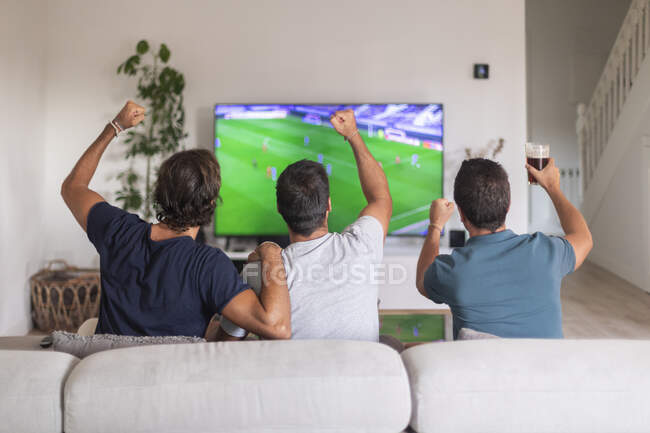 Tres amigos viendo un partido de fútbol en casa bebiendo cerveza - foto de stock
