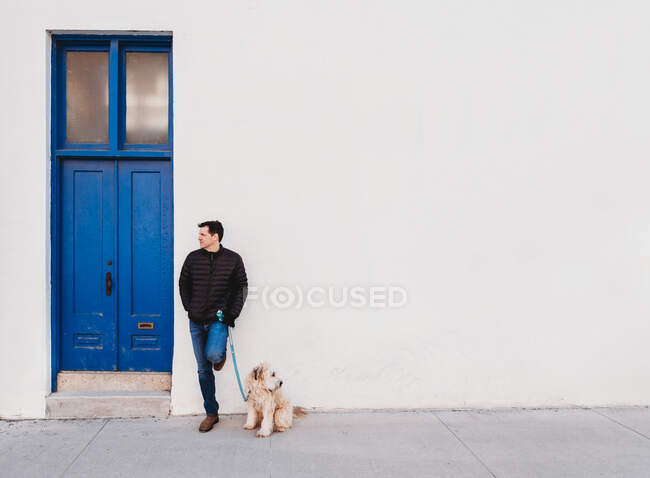 Uomo e cane in piedi da soli accanto a una porta blu di un edificio bianco. — Foto stock