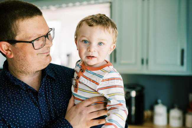 Отец держит своего маленького сына улыбаясь ему стоя на кухне — стоковое фото