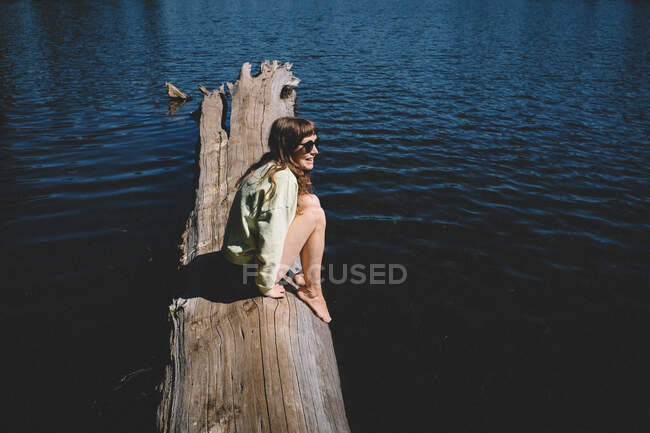 Niedliche Brünette mit Bangs, Sonnenbrille sitzt auf einem Baumstamm, umgeben von Wasser — Stockfoto