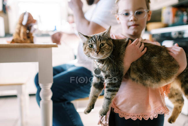Pequena menina criança segurando seu gato de estimação bonito em seus braços na cozinha — Fotografia de Stock