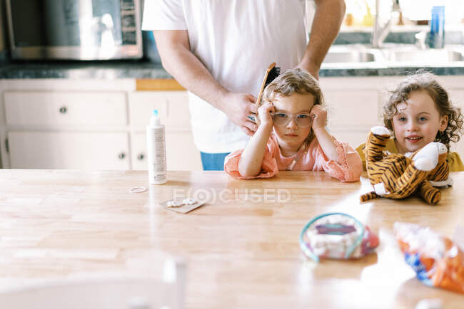 Uma menina recebendo seu cabelo feito por seu pai na mesa da cozinha — Fotografia de Stock