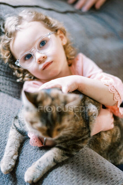 Una niña con gafas acariciando a su amigo gato en el sofá - foto de stock