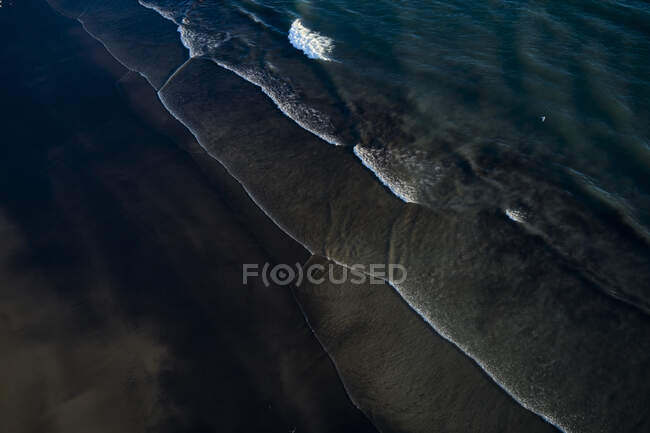 Вид с воздуха на прозрачные океанские волны, идущие по темной холодной береговой линии с белой пеной — стоковое фото